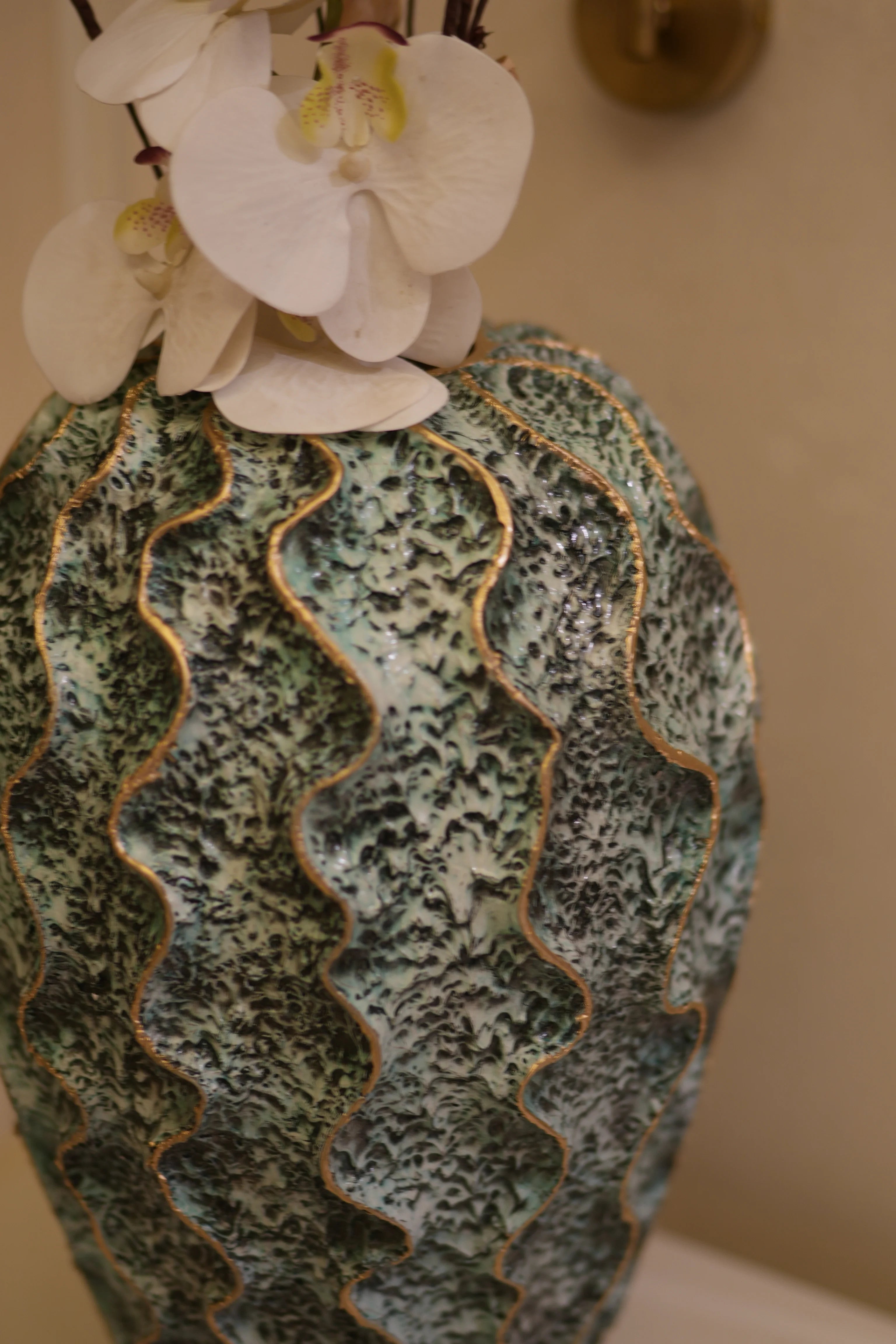 A close-up of a decorahaus resin vase with a textured, wavy pattern in green, blue, and black, accented with gold-colored trim, next to white orchid flowers.
