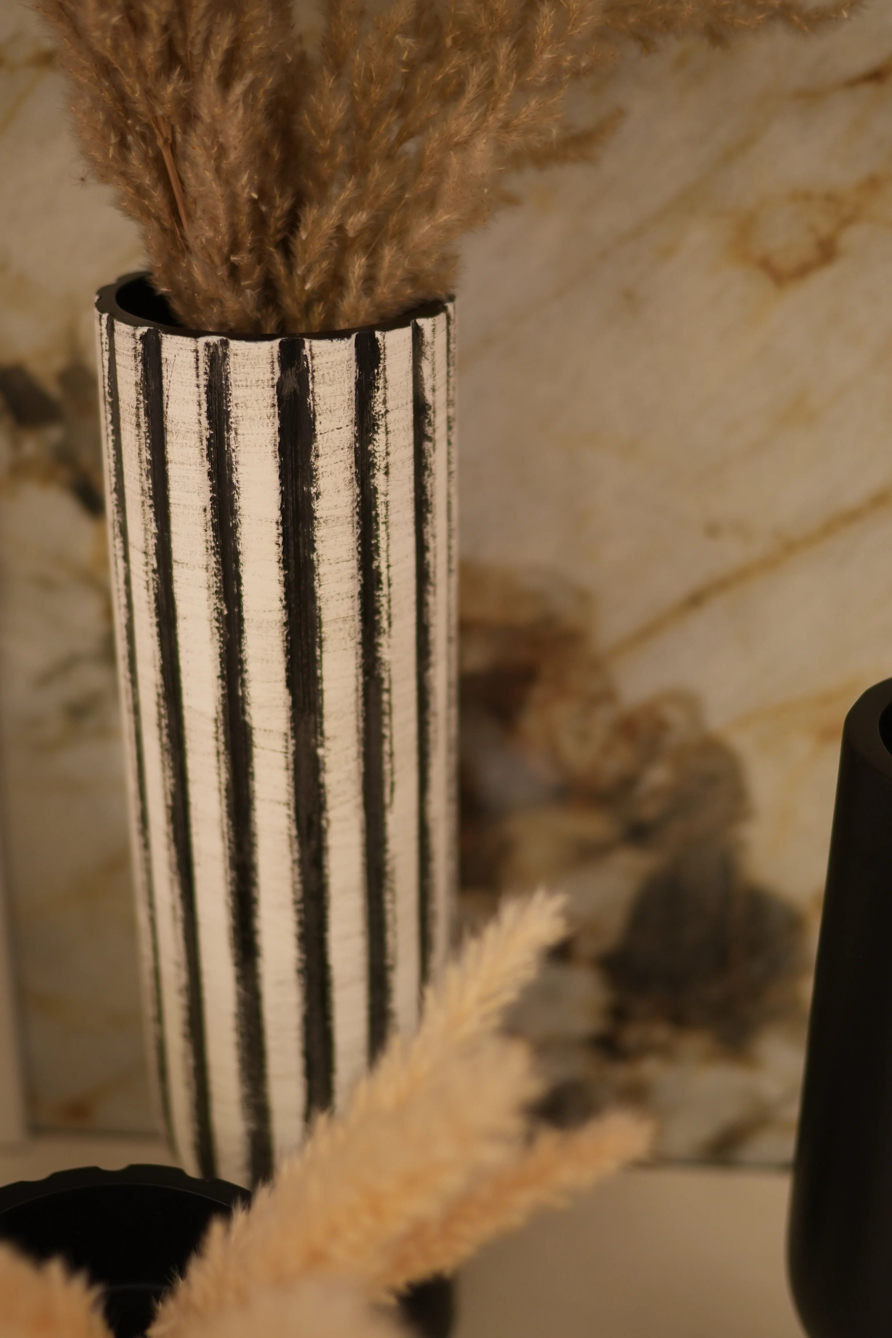 A close-up of a cylindrical decorative object with alternating black and white horizontal stripes, featuring fluffy beige and brown feather or faux-fur elements extending from both ends, set against a marble-patterned background.