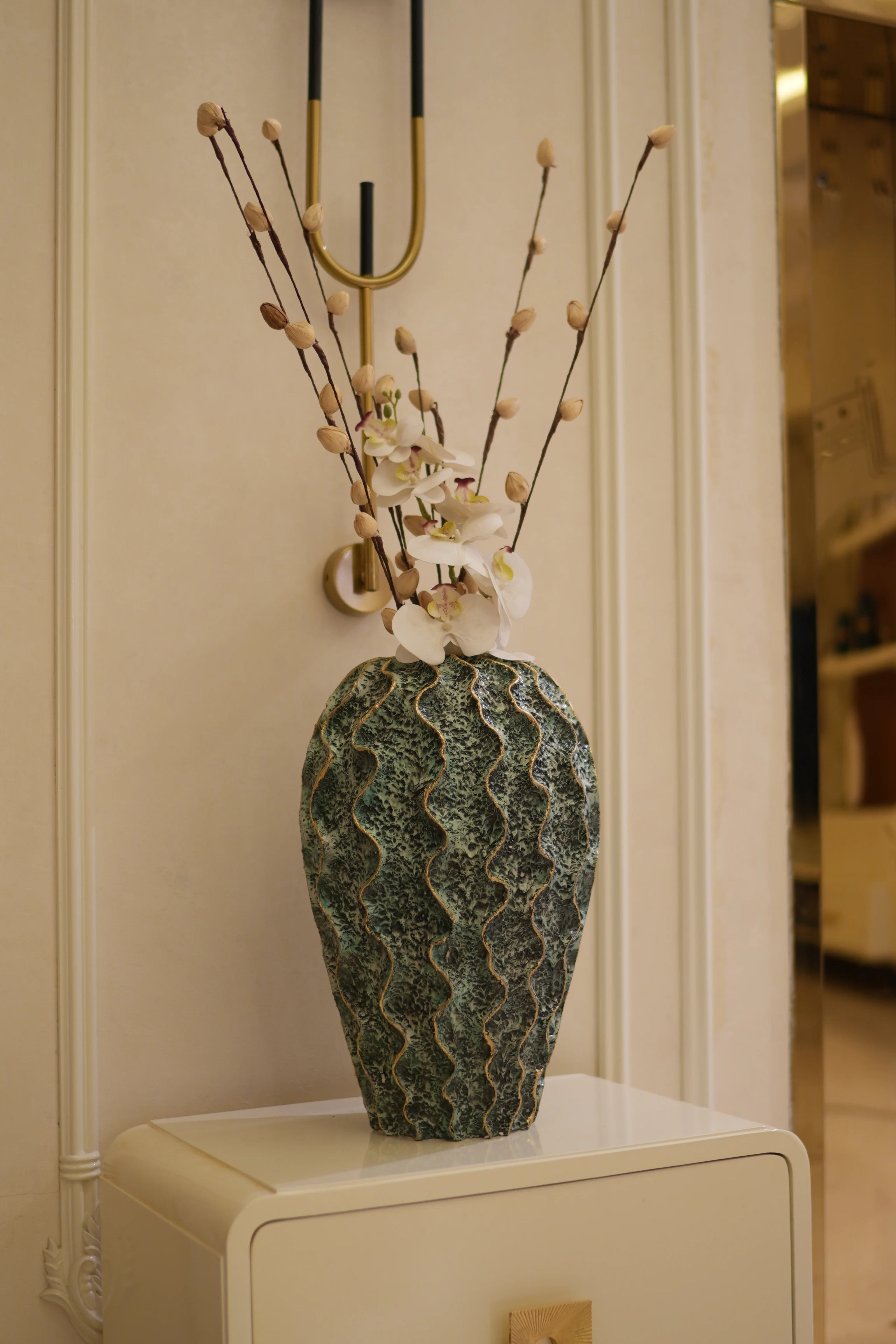 A decorative Decorahaus resin vase with a textured, wavy green and blue glaze, holding dried branches with small white flowers, displayed on a cream-colored mantelpiece.