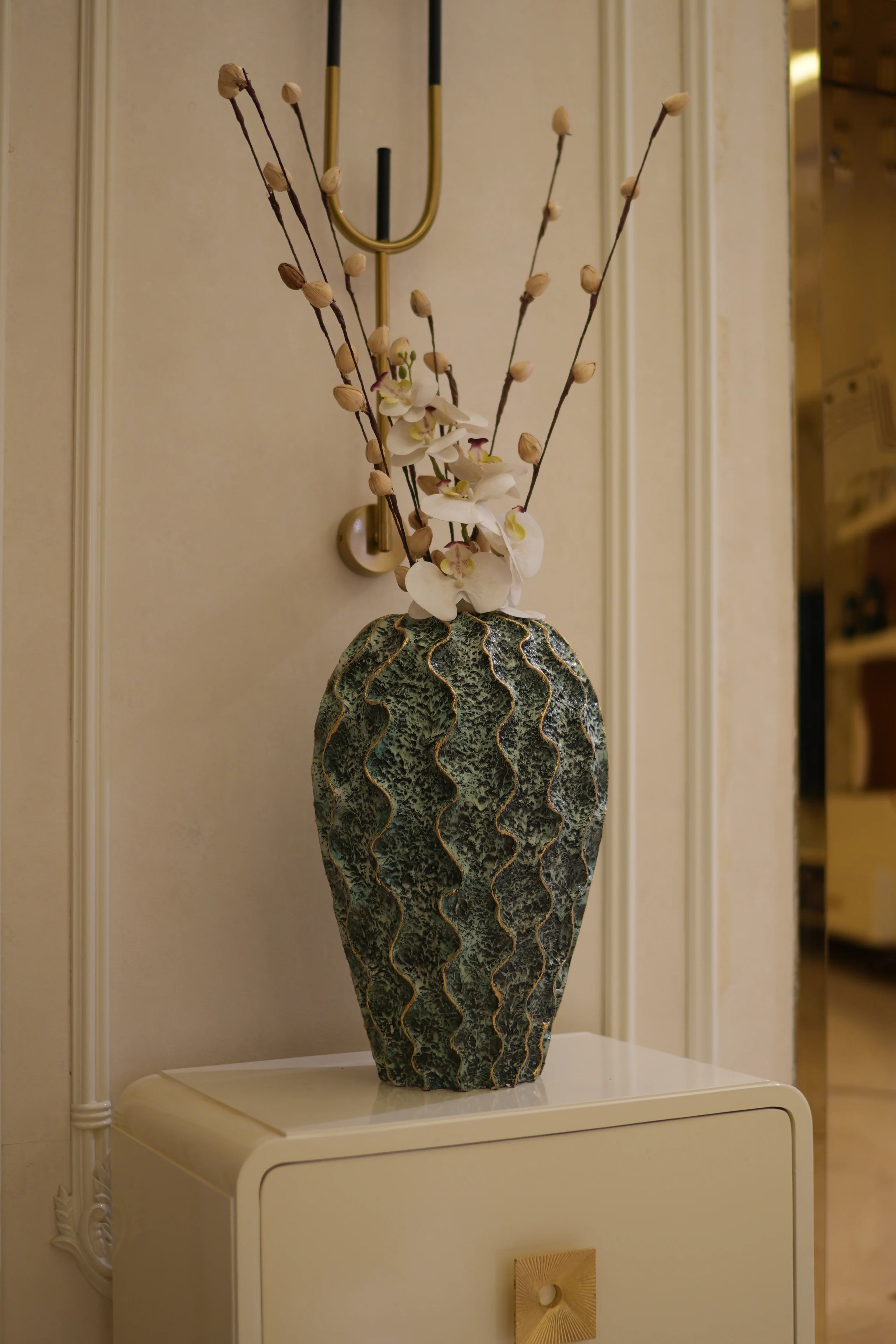 A decorative decorahaus resin vase with a textured, wavy green and blue pattern, holding dried branches with small white flowers, set against a warm beige background.