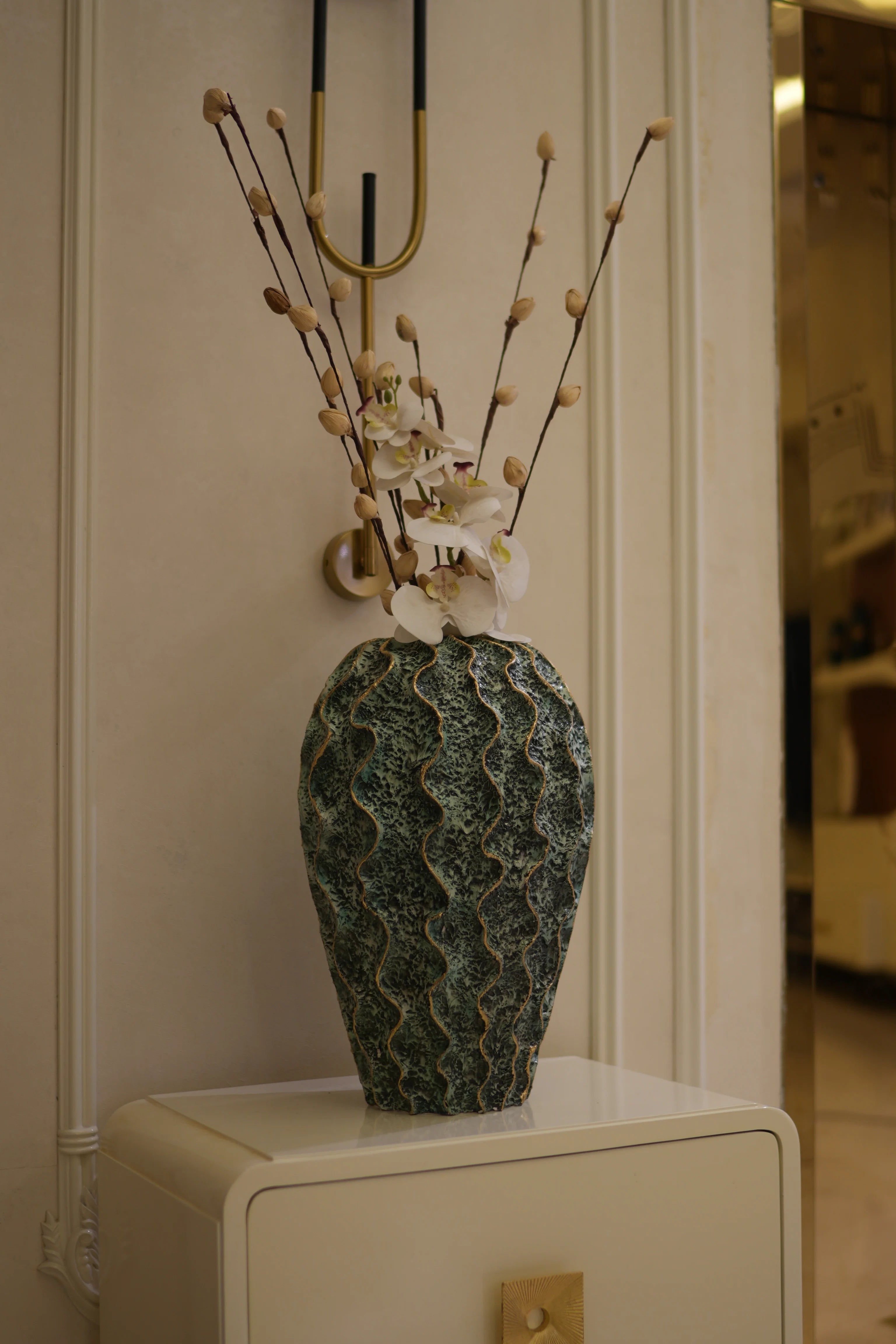 A decorative textured resin vase with a wavy pattern in dark green and gray, holding dried branches with small cream-colored buds, displayed on a white mantelpiece next to a cream-colored cabinet.