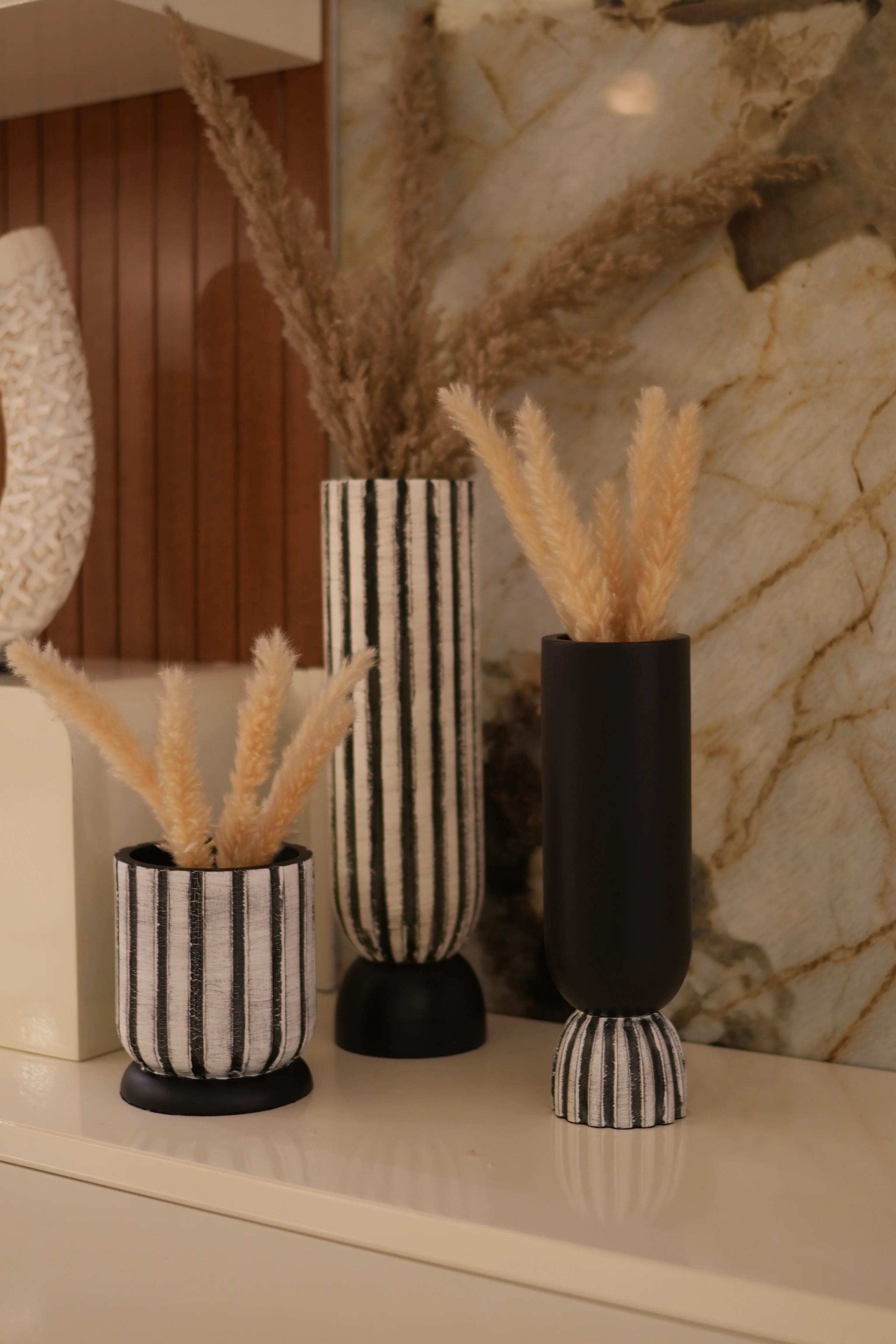 Three decorative pillar candle holders with black and white striped fabric wraps and beige dried grass accents, arranged on a marble surface next to a wooden floor.