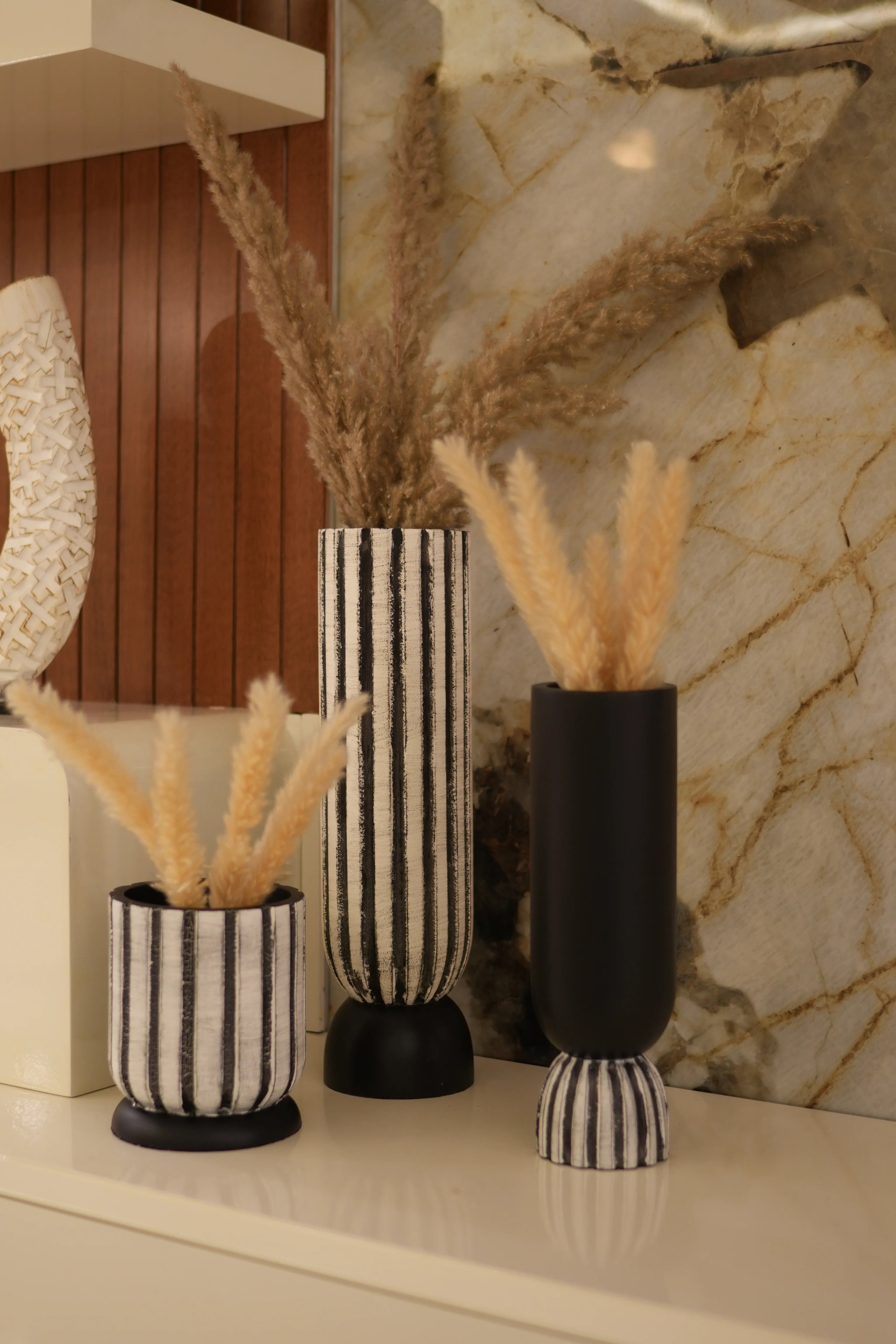 Three decorative decorahaus black and white striped fabric-covered vases with dried beige grass stems, arranged on a marble surface next to a wooden floor.