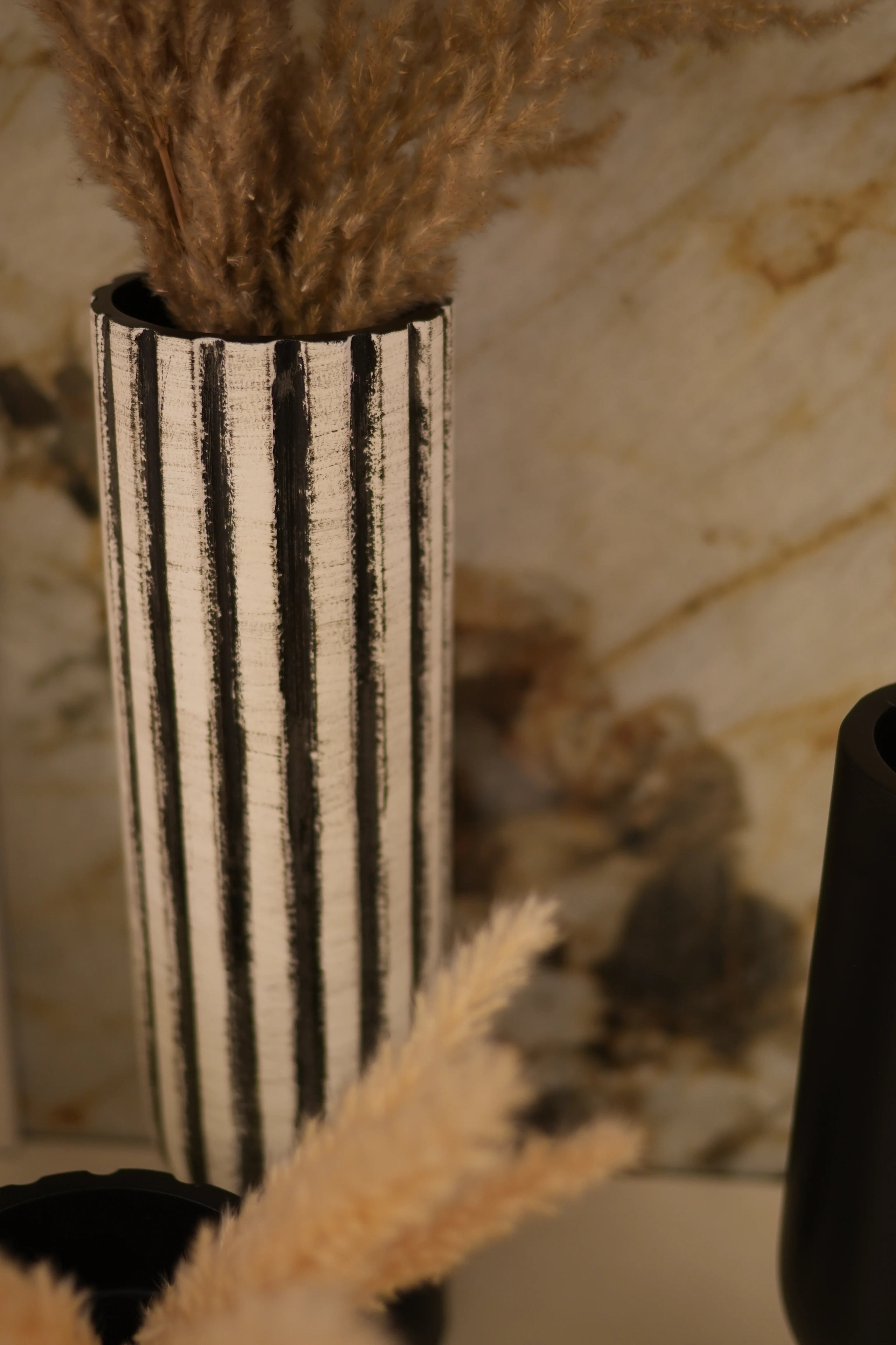 A close-up of a decorahaus cylindrical decorative object with alternating black and off-white horizontal stripes, partially surrounded by fluffy beige and brown textured materials on a light marble-like surface.